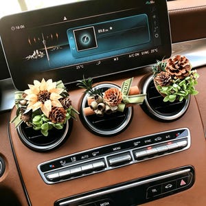 May include: Three car air vent decorations with floral arrangements. Each vent clip features a different design, including pinecones, dried flowers, and cinnamon sticks, accented with green ribbon. The car's dashboard is brown.