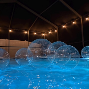 May include: Multiple large, transparent, spherical inflatable balls floating on the surface of a swimming pool at night. The water is illuminated with a vibrant blue light, and the clear spheres feature a subtle, textured surface pattern.