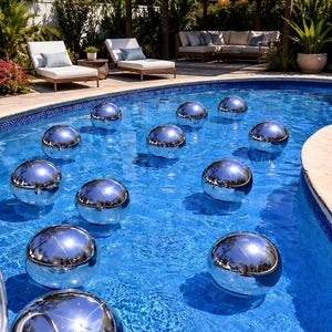 May include: A collection of twelve spherical, polished silver-colored floating pool ornaments. These smooth, reflective globes are distributed across the surface of a bright blue swimming pool, creating a mirrored effect of the surrounding outdoor environment.