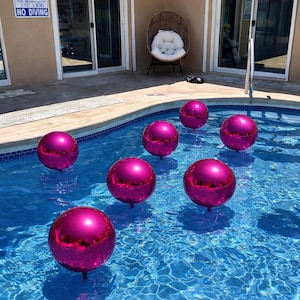 May include: A swimming pool filled with clear blue water, featuring several large, spherical, magenta-colored balloons floating on the surface. The pool is surrounded by a patio and a building with sliding glass doors.