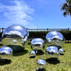 May include: A collection of seven spherical, silver, reflective balloons of varying sizes arranged on a green lawn. The metallic surfaces of the balloons mirror the surrounding outdoor environment, including the blue sky and nearby greenery.