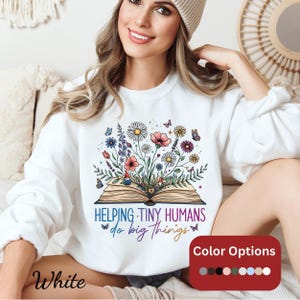Puede incluir: Sudadera blanca con un diseño de libro floral. El diseño presenta un libro abierto con flores y mariposas coloridas, y el texto "Helping Tiny Humans do big things". Las opciones de color se muestran en un recuadro rojo.