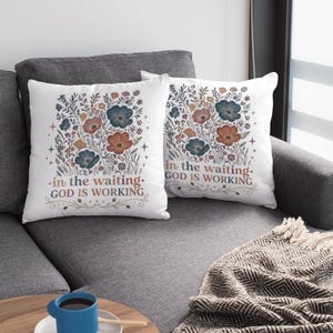 Pode incluir: Duas almofadas brancas com desenhos florais em tons de azul, laranja e castanho. As almofadas têm o texto "in the waiting. GOD IS WORKING". Estão num sofá cinzento.