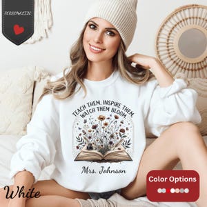 Op de afbeelding: Witte sweatshirt met de tekst "TEACH THEM, INSPIRE THEM, WATCH THEM BLOOM" en "Mrs. Johnson". Het ontwerp toont een open boek met bloemen en vlinders. Kleuropties worden weergegeven in een rode doos.
