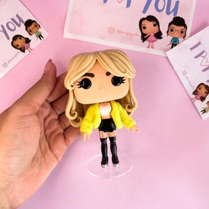 May include: A vinyl collectible figure with blonde hair, wearing a bright yellow fuzzy jacket, a white crop top, black shorts, and black thigh-high boots. The figure stands on a clear circular base and features large black eyes and a small nose.