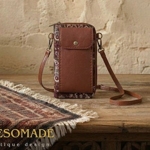 May include: A brown leather crossbody bag with a patterned fabric accent. The bag has a flap closure with a button and a long, adjustable strap. The bag is sitting on a wooden surface. The text "MESOMADE Antique design" is visible.