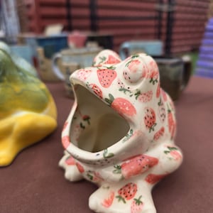May include: A ceramic frog-shaped planter or container with a wide, open mouth. The exterior is cream-colored and decorated with a repeating pattern of small red strawberries and green leaves. The frog has two prominent eyes on top of its head.