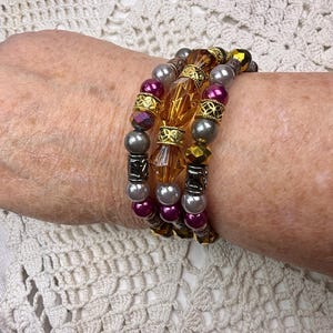 May include: A multi-strand beaded bracelet featuring a combination of amber-colored faceted crystal beads, metallic gold spacers, polished grey pearls, and deep magenta round beads arranged in a layered design with dark metal accents.
