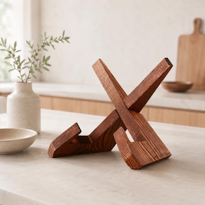 May include: A wooden cookbook stand in a warm brown finish. The stand is constructed with intersecting angled pieces, creating an X-shaped design. The stand is designed to hold a cookbook or tablet. The stand is on a light-colored countertop.