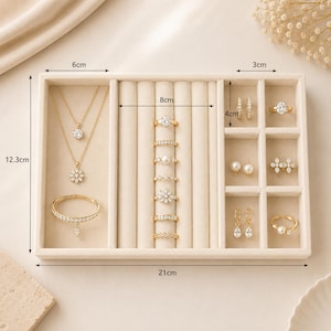 May include: A beige velvet jewelry organizer tray measuring 21 cm by 12.3 cm, featuring compartments for necklaces, a bracelet, seven rings, and six pairs of earrings. The tray includes labeled dimensions of 6 cm, 8 cm, 3 cm, and 4 cm for its sections.