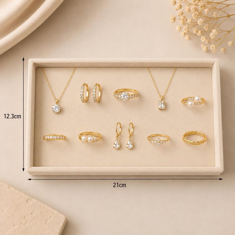 May include: A collection of gold-toned jewelry including two necklaces, three pairs of earrings, and six rings arranged on a rectangular tray measuring 12.3 cm by 21 cm. The pieces feature clear stones and white pearls in various settings.