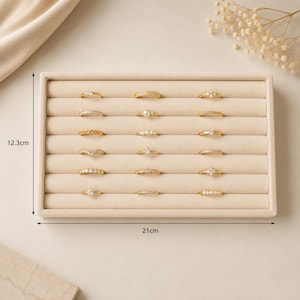 May include: A rectangular beige velvet ring display tray featuring six horizontal slots holding eighteen gold-toned rings with various stones and pearls. The tray measures 12.3 inches by 21 inches as indicated by the black dimension lines.