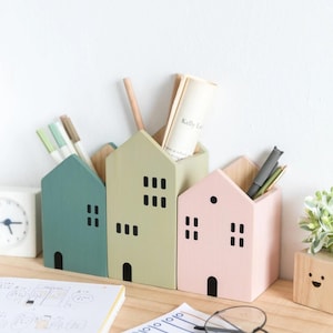 May include: Three wooden house-shaped desk organizers in pastel colors: teal, green, and pink. Each has a black door and window cutouts. The organizers hold pens, pencils, and small notebooks, sitting on a wooden desk.