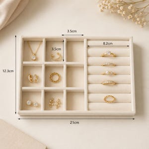 May include: A beige velvet jewelry organizer tray measuring 12.3 cm by 21 cm, featuring multiple compartments and ring slots. It contains gold-toned jewelry including a necklace, earrings, a cuff, and five rings with various stone settings.