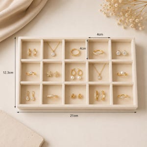 May include: A beige velvet jewelry organizer tray with fifteen compartments containing various gold-toned rings, earrings, and necklaces. The tray measures 21 cm by 12.3 cm, with individual compartments measuring 4 cm by 4 cm, as indicated by labels.