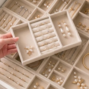 May include: A beige velvet jewelry organizer tray featuring multiple compartments for rings, earrings, and necklaces. The tray contains various gold-toned jewelry pieces with white pearls and clear stones, including a floral brooch and drop earrings.