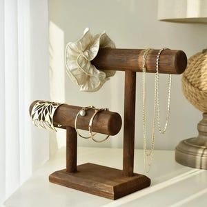 May include: A dark brown wooden jewellery display stand with two horizontal bars. A gold bracelet, gold hoop earrings, and a white scrunchie are displayed. Two gold necklaces hang from the top bar.