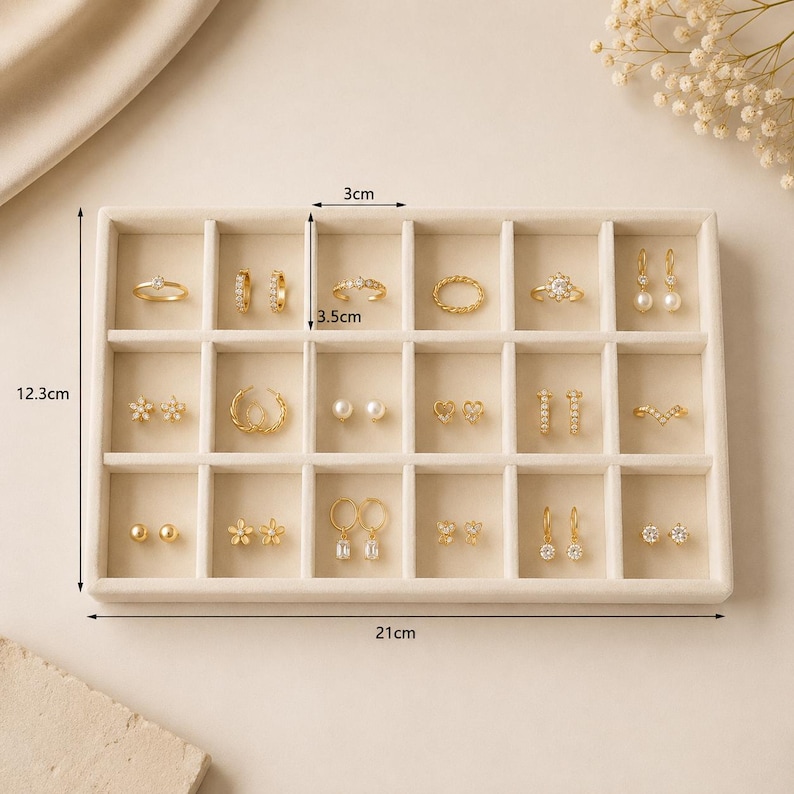 May include: A beige velvet jewelry organizer tray with 15 compartments containing various gold-toned rings and earrings. The tray measures 21 cm by 12.3 cm, with individual slots measuring 3 cm by 3.5 cm, showcasing a collection of jewelry pieces.