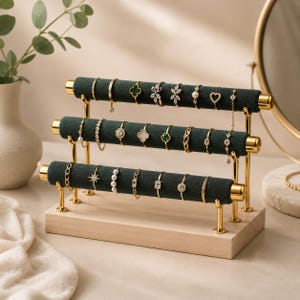 May include: A three-tier jewelry display stand featuring dark green velvet-covered bars supported by gold-toned metal rods on a light wood base. The stand holds various gold-toned rings and a delicate chain bracelet arranged across the three levels.