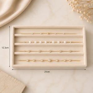 May include: Four gold-tone chain bracelets displayed on a beige velvet jewelry tray. The tray measures 12.3 cm by 21 cm. The bracelets feature various accents including small clear stones, white pearls, and flower-shaped charms along the chains.