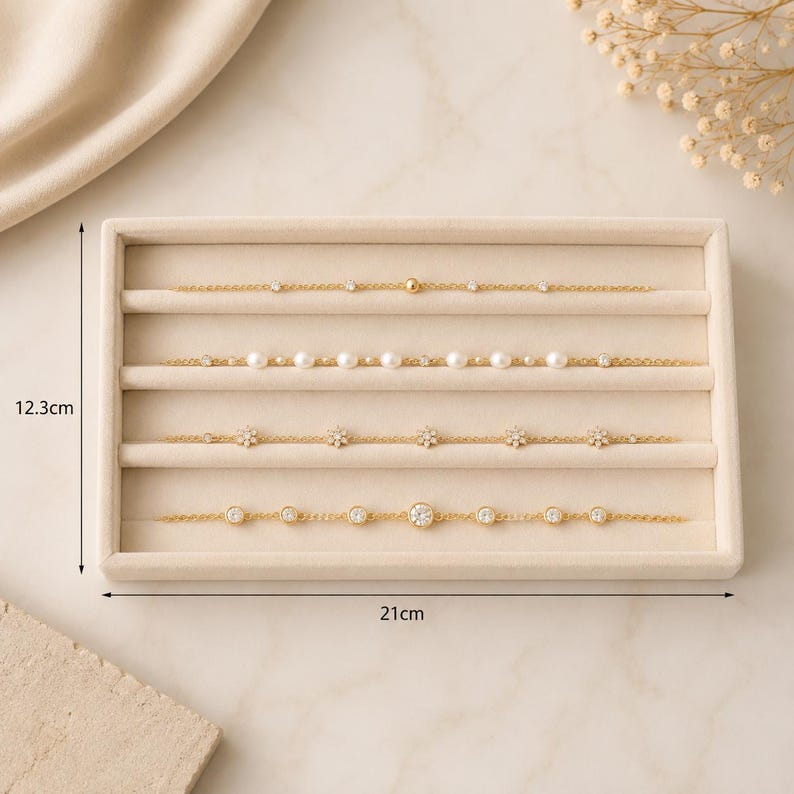 May include: Four gold-tone chain bracelets displayed on a beige velvet jewelry tray. The tray measures 12.3 cm by 21 cm. The bracelets feature various accents including small clear stones, white pearls, and flower-shaped charms along the chains.
