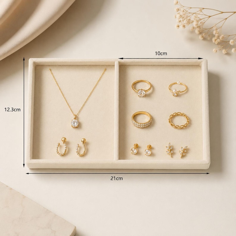 May include: A beige jewelry organizer tray measuring 21 cm by 12.3 cm with a 10 cm wide right compartment. It holds a gold necklace with a clear stone pendant, two horseshoe earrings, four gold rings, and two pairs of small gold stud earrings.