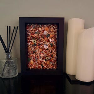 May include: A framed artwork filled with various seashells in shades of brown, orange, and white. Beside it are two white pillar candles and a decorative glass diffuser with black reeds.
