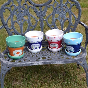 May include: Four small ceramic flower pots with matching saucers, each painted in a different color with a fruit theme label. The pots are labeled Orange Marmalade, Grape Jelly, Strawberry Jam, and Blueberry Jam, featuring small painted fruit.