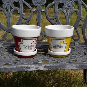May include: Two ceramic plant pots with matching saucers. One pot is red with a white rim and a white label graphic reading TOMATO KETCHUP. The other pot is yellow with a white rim and a white label graphic reading YELLOW MUSTARD.
