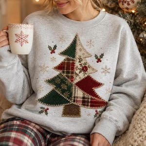 May include: A light grey crewneck sweatshirt featuring a large patchwork-style Christmas tree graphic composed of various green, red, and cream plaid and floral fabric patterns. The design includes small holly berries and snowflakes.
