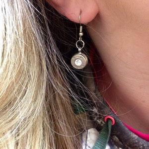 May include: Silver bullet earrings with a clear rhinestone in the center. The earrings are hanging from a person's ear.