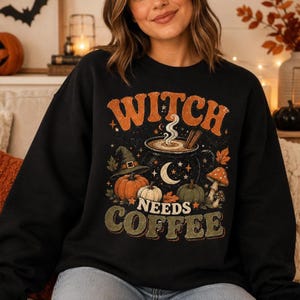 May include: A black long-sleeved sweatshirt featuring a graphic print with the text "WITCH NEEDS COFFEE". The design includes a steaming cauldron, a witch hat, pumpkins, mushrooms, a crescent moon, and stars in orange, green, and white colours.