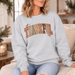 May include: A light grey crewneck sweatshirt featuring the word THANKFUL printed across the chest in large block letters. Each letter displays a unique autumn-inspired pattern, including plaid, floral, and wavy line designs in shades of brown and tan.