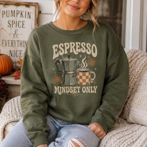 May include: An olive green crewneck sweatshirt featuring a graphic print with the text "ESPRESSO MINDSET ONLY". The design includes a silver moka pot, a steaming mug with a brown and white checkered pattern, and small star and sunburst accents.