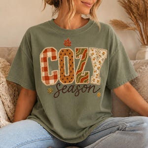 May include: An olive green t-shirt featuring the text COZY in large block letters with varied patterns including red plaid, brown polka dots, green wavy lines, and yellow floral print. The word season is written below in cursive script.