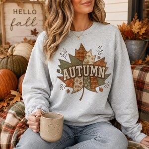 May include: A light grey crewneck sweatshirt featuring a large central graphic of a maple leaf composed of various brown, green, and tan plaid and floral patterns. The word AUTUMN is printed in bold, light-coloured block letters across the leaf.