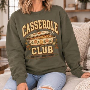 May include: An olive green crewneck sweatshirt featuring a graphic of a casserole dish with floral patterns. The text reads CASSEROLE CLUB, HOT DISHES • GOOD COMPANY, and EST. THANKSGIVING. The design includes a spoon and small star accents.