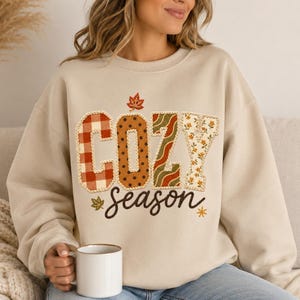 May include: A beige crewneck sweatshirt featuring the word COZY in large, multi-patterned block letters with a red plaid, brown polka dot, green wavy, and floral print. The word season is written below in brown script with small leaf accents.