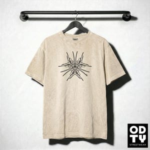 May include: A beige t-shirt with a black abstract design hanging on a black hanger. The t-shirt has a crew neck and short sleeves. The logo "OD TY STREETWEAR" is in the bottom right corner.