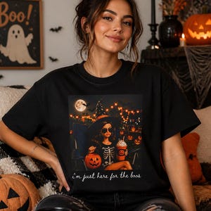 Può includere: Una t-shirt nera a girocollo con una stampa grafica di una persona che indossa un cappello da strega e una maglietta con scheletro, che tiene in mano un secchiello a forma di zucca e una bevanda. Il testo "i'm just here for the boos" è in bianco.