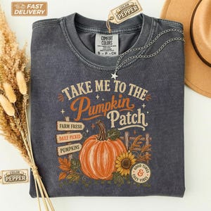 Può includere: Una t-shirt girocollo grigio antracite con una grafica raffigurante una grande zucca arancione, un girasole e insegne in legno con le scritte "Take Me To The Pumpkin Patch", "Farm Fresh", "Daily Picked" e "Pumpkins" con un piccolo logo.