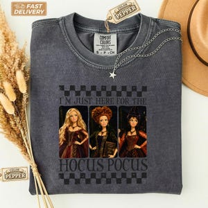 Può includere: Una t-shirt girocollo grigio antracite con una stampa grafica di tre bambole vestite con abiti scuri, posizionate tra un bordo a scacchi. Il testo recita I'M JUST HERE FOR THE HOCUS POCUS. L'etichetta riporta la scritta COMFORT COLORS.
