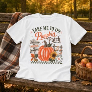 Può includere: Una t-shirt bianca a maniche corte con una stampa grafica recante il testo Take Me To The Pumpkin Patch. Il design include una grande zucca arancione, un cartello in legno, un girasole e un bordo a scacchi bianchi e neri.
