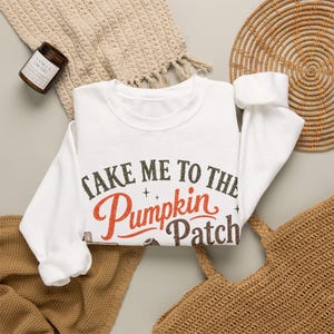 Può includere: Una felpa bianca a maniche lunghe con girocollo che riporta la scritta "TAKE ME TO THE Pumpkin Patch" in lettere marrone scuro e arancioni. Il design include piccoli accenti a forma di stella e un grafico di una zucca in basso.