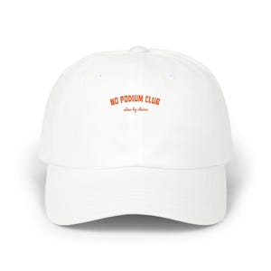 May include: A white baseball cap featuring the text NO PODIUM CLUB in a bold orange font, with the phrase slow by choice written underneath in a smaller, cursive orange script. The cap has a structured crown and a curved brim.