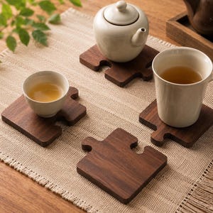Op de afbeelding: Een theeset met een witte theepot en theekopjes op houten puzzelvormige onderzetters. De onderzetters zijn donkerbruin en liggen op een beige tafelkleed. De kopjes zijn gevuld met thee.