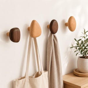 Peut inclure: Quatre patères en bois fixées au mur, de formes et de teintes variées, allant du noyer foncé au chêne clair. Les patères sont fixées sur un mur uni, avec un sac fourre-tout en toile beige suspendu à l'une et une écharpe en tissu beige clair à une autre.