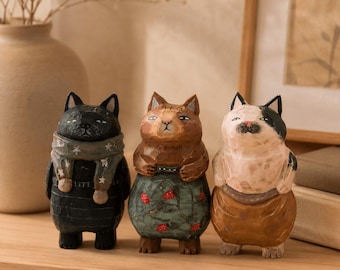 Puede incluir: Tres figuras de gato talladas en madera a mano dispuestas en fila. El gato de la izquierda es negro con una bufanda gris con estrellas blancas. El gato central es marrón con una falda verde azulado con fresas rojas. El gato derecho es blanco y negro.