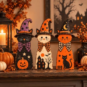 Op de afbeelding: Drie houten kattenbeeldjes versierd voor Halloween. De linker kat is zwart met een paarse hoed en pompoen. De middelste kat is wit met een oranje hoed en spookhuis. De rechter kat is oranje met een zwarte hoed en silhouet van een zwarte kat.