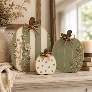 May include: Three flat wooden pumpkin decorations in sage green and cream. The largest features vertical stripes and a floral relief, the medium has a solid green finish with floral relief, and the smallest is cream with green polka dots.